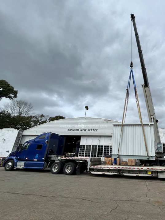 Flatbed truck and crane preparing to load UPS enclosure at shipping facility
